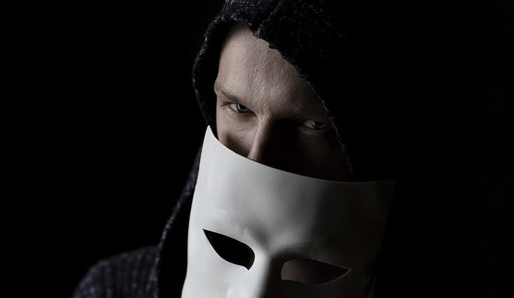 A shadowy figure with a glint in his eye removes his mask.