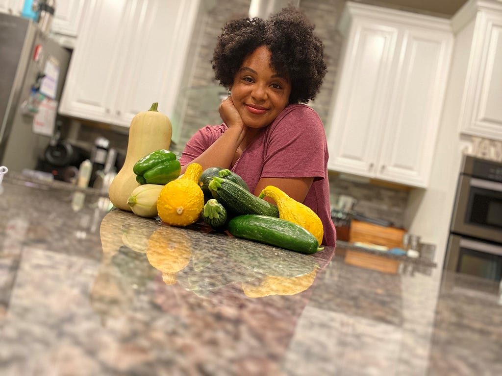 Woman in kitchen with butternut squash, green pepper, yellow squash, zucchini and cucumber.