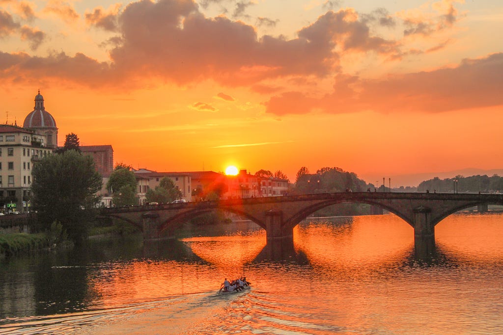Bridge in Florence over Arno River with rowers under glow of sunset of bright orange and pink sky