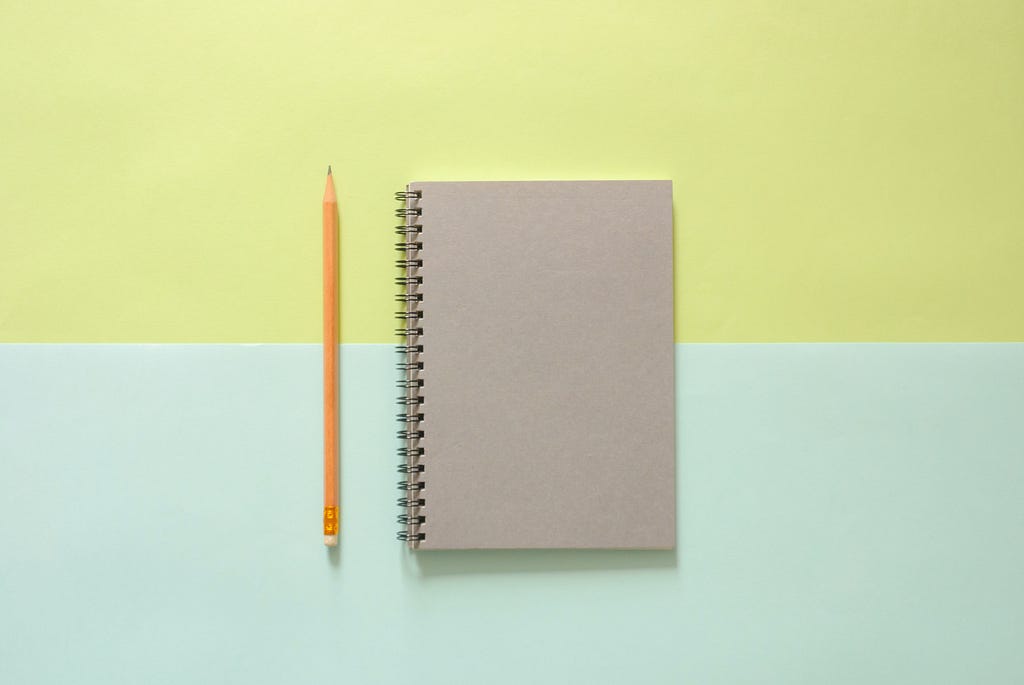 A yellow pencil and spiral notebook are laid side by side.