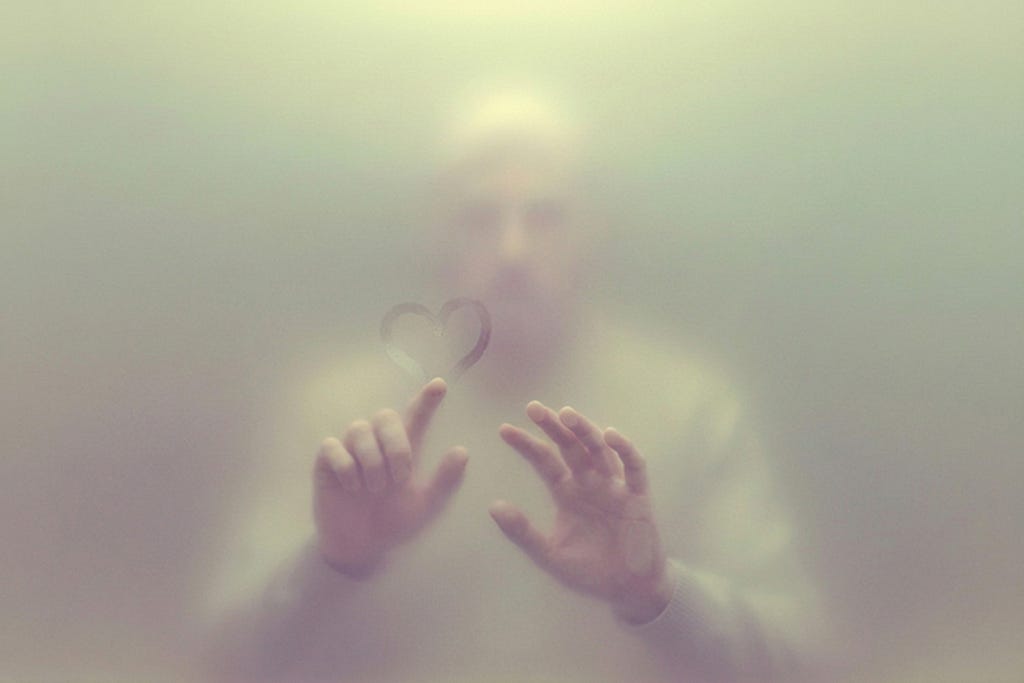 A heart shape drawn on a foggy glass barrier with hands visible below, representing affection directed toward one’s own confinement.