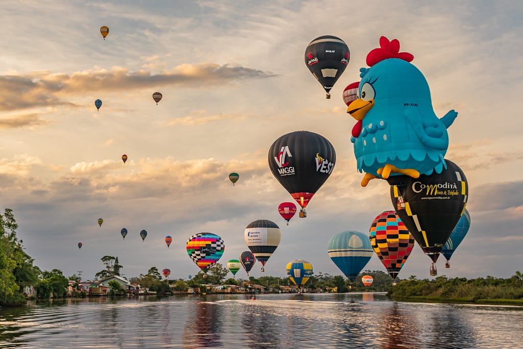 Foto: Festival de Balões da cidade de Torres. O céu está claro com algumas nuvens. Sobre a água cerca de 24 balões voam, coloridos, e chamando a atenção mais a frente na imagem um enorme balão no formato da Galinha Pintadinha em azul.