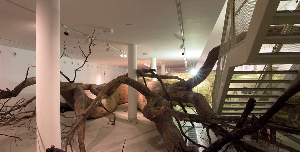 Instalação de troncos e raízes de árvore em escala ampliada dentro de um salão branco, perpassando duas colunas e uma escada lateral.