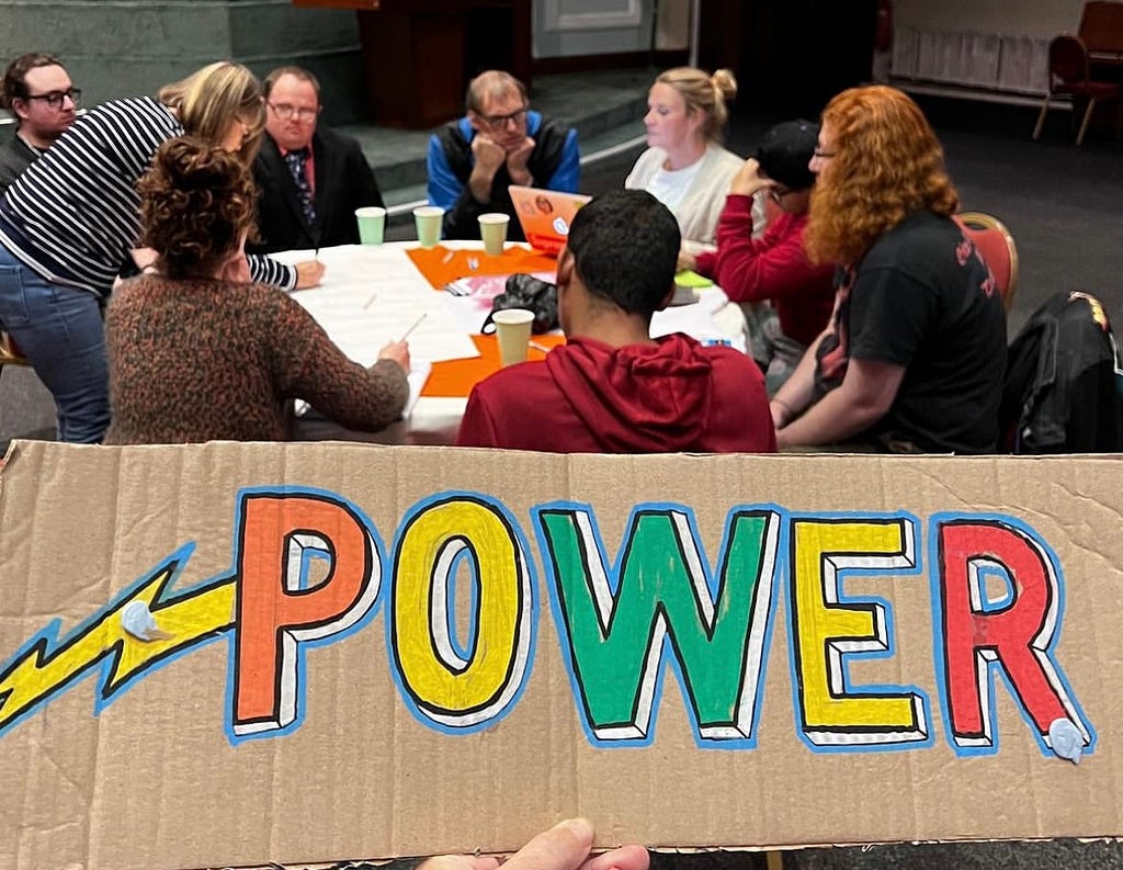 A group of people sit round a table engaged in discussion. In the front of the image is a sign that says “power”.