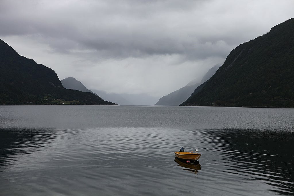 A mountain fjord with a yellow boat on calm water. The mountains narrow to an opening in the middle.