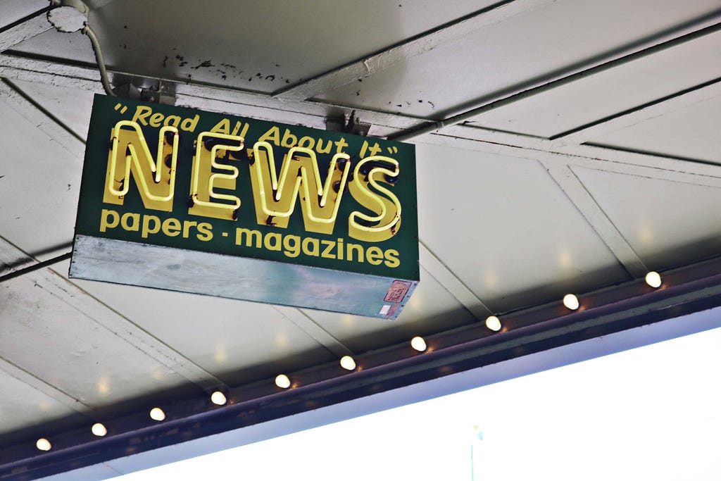 A neon sign hanging beneath an illuminated marquee that says: “Read All About It” (newline) News (newline) papers * magazines.