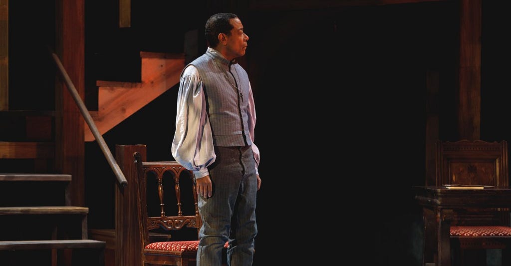 A photograph of a man with brown skin and short dark hair, looking off-camera to the right. He wears a light blue high-necked vest and a lighter blue shirt with billowing sleeves over gray pants, and stands in front of a wooden staircase.