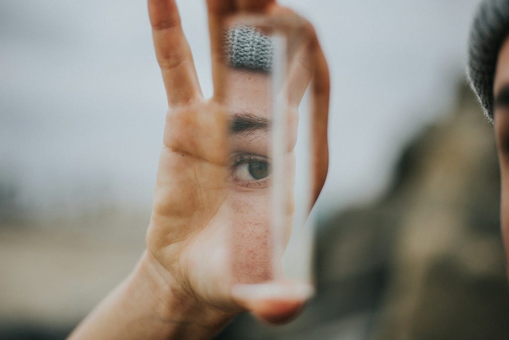 A woman holds up a shard of mirror, showing the reflection of her eye held in the palm of her hand