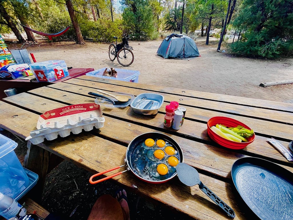 Eggs in a pan and other breakfast supplies on a picnic table, with a bicycle, tent and hammock in the background.