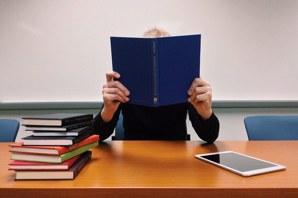 Guy holding up a book to his face with both hands, ironically, an unlikely studying pose, with a stack of books to his right