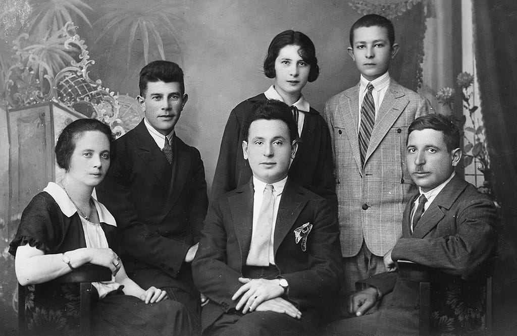 A group of young adults poses in front of a painted formal backdrop. The four men are wearing suits and ties. The two women are wearing blouses with white collars. All look at the camera.