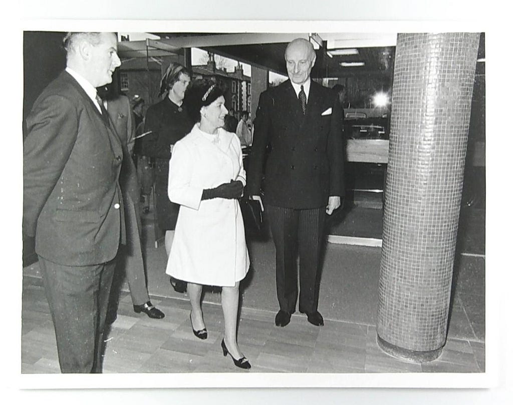 Black and white photograph showing Princess Margaret arriving at the Kennedy Institute. She is in the centre of the image with two men either side and some people behind her.