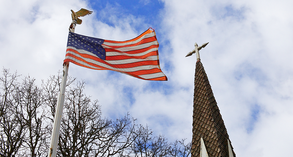 A U.S. flag hangs on an eagle-topped flagpole against a blue sky. To its right is a church steeple, nearly equal in height.