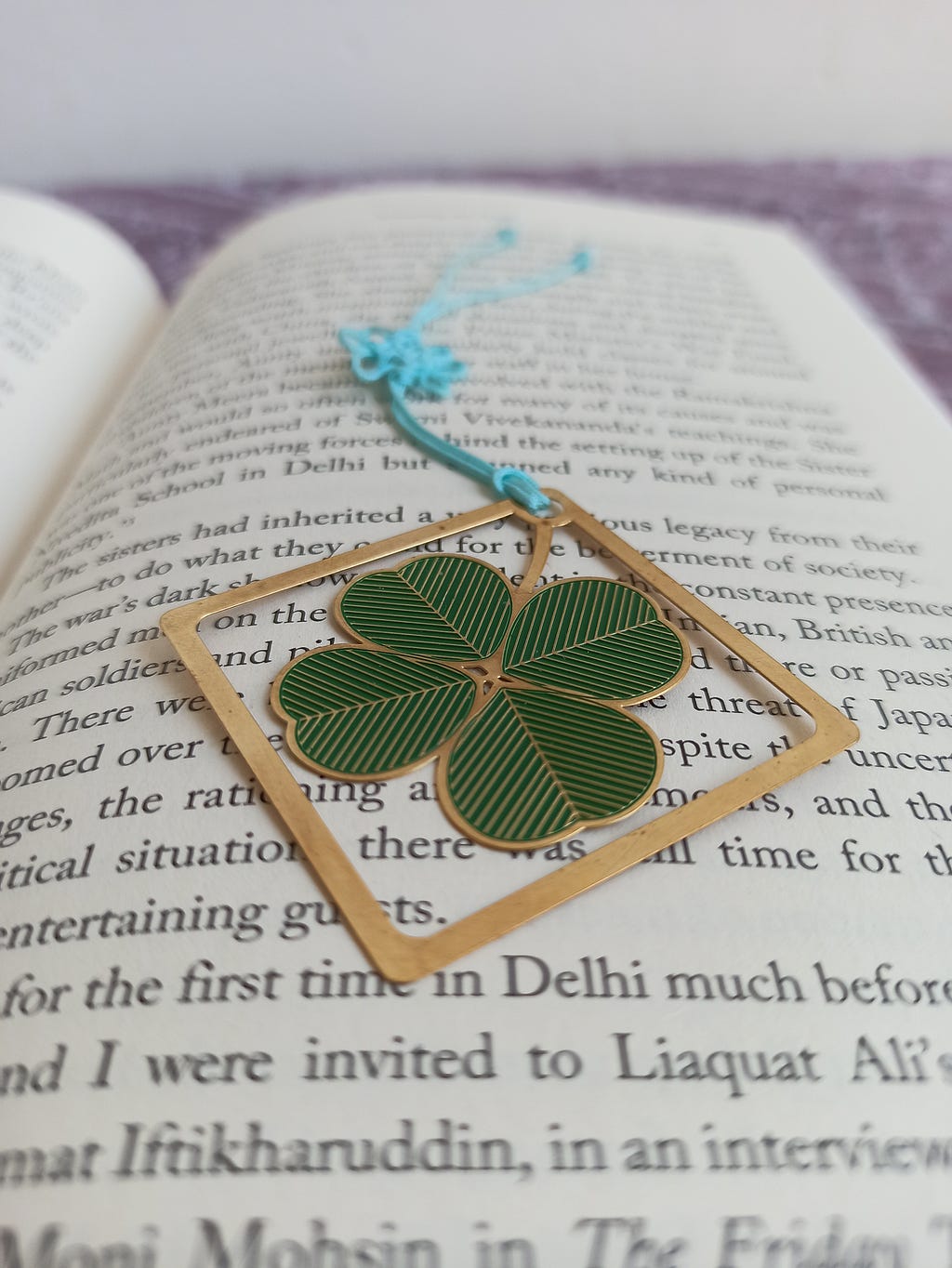 The image shows an open book. The words are blurred. On top of the page is a bookmark with a four leave clover pattern on it.