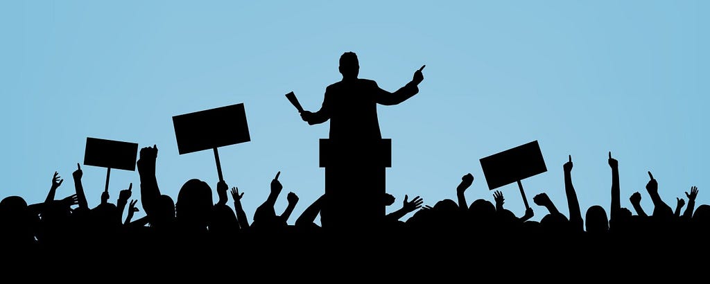 In black silhouettes, a speaker stands at a podium in front of a crowd raising its hands and holding up placards.