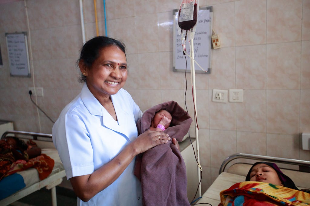 A health worker smiles as she holds a newborn baby while standing next to a an IV pole and a hospital bed occupied by a woman.