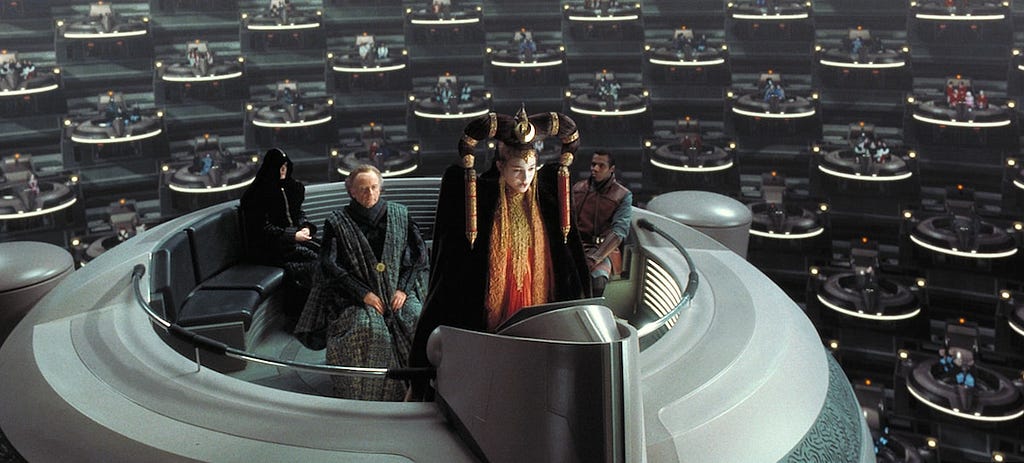 Queen Amidala dolled up to the nines, addressing the Galactic Senate to protest the Trade Federation’s invasion of Naboo. She wears an elaborate headdress, quasi-Kabuki makeup and a dramatic ceremonial outfit. She is standing at a podium on a floating space pod, surrounded by a group of rather bored-looking men.