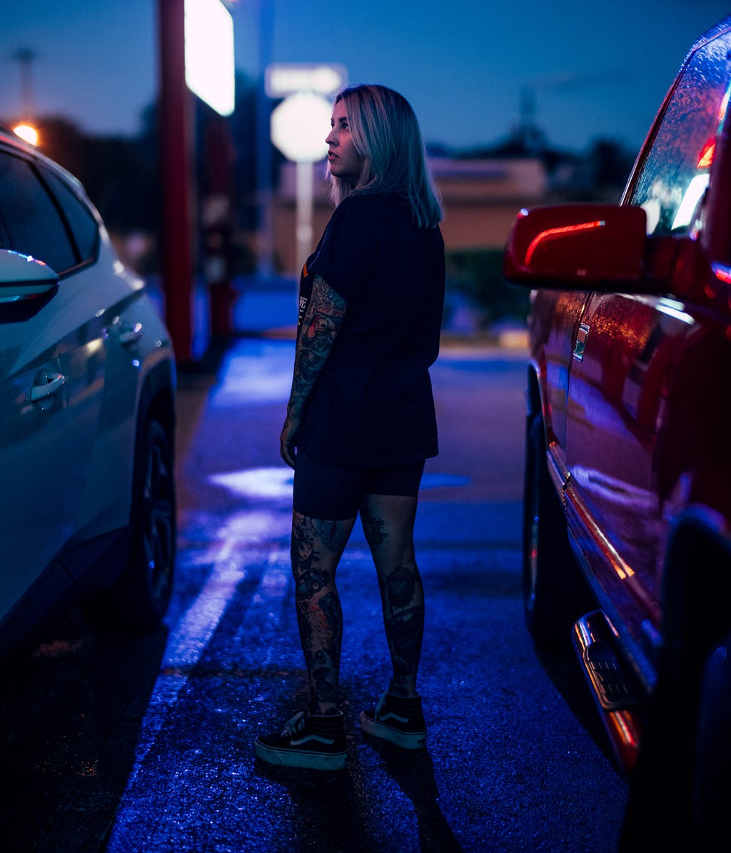 Erin stands in a dimly lit parking lot between two cars, light flaring in red and blue across the concrete — a moment suspended between day and night.