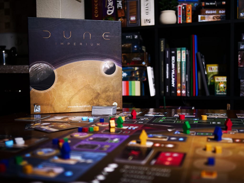 Photo of Dune Imperium’s box with the game set up on a table. Behind it is a shelf with several other board games and table top RPG books.