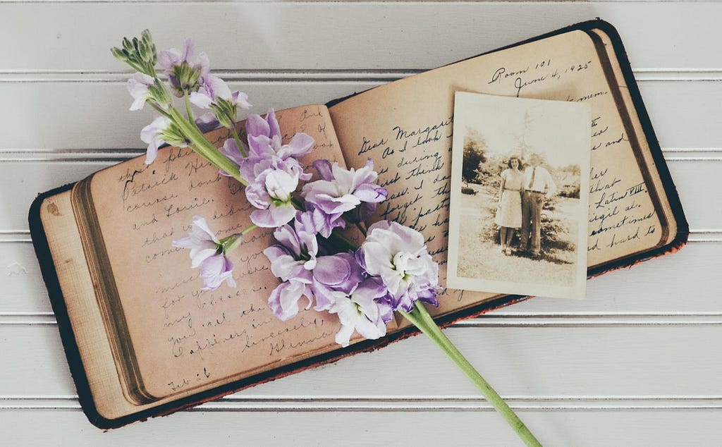 An old handwritten diary lies open on a white background with a a black and white photograph and some flowers on top of it