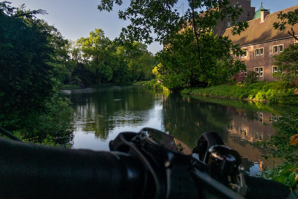 View of the pond that surrounds the water castle Wittringen with parts of the handlebars of my bike in the foreground. The castle offers a decent spot for a slightly longer ride. Gladbeck, Germany, May 26, 2023.