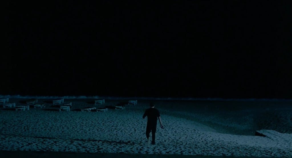 Homem vestido de roupas pretas andando em direção ao mar numa praia à noite.
