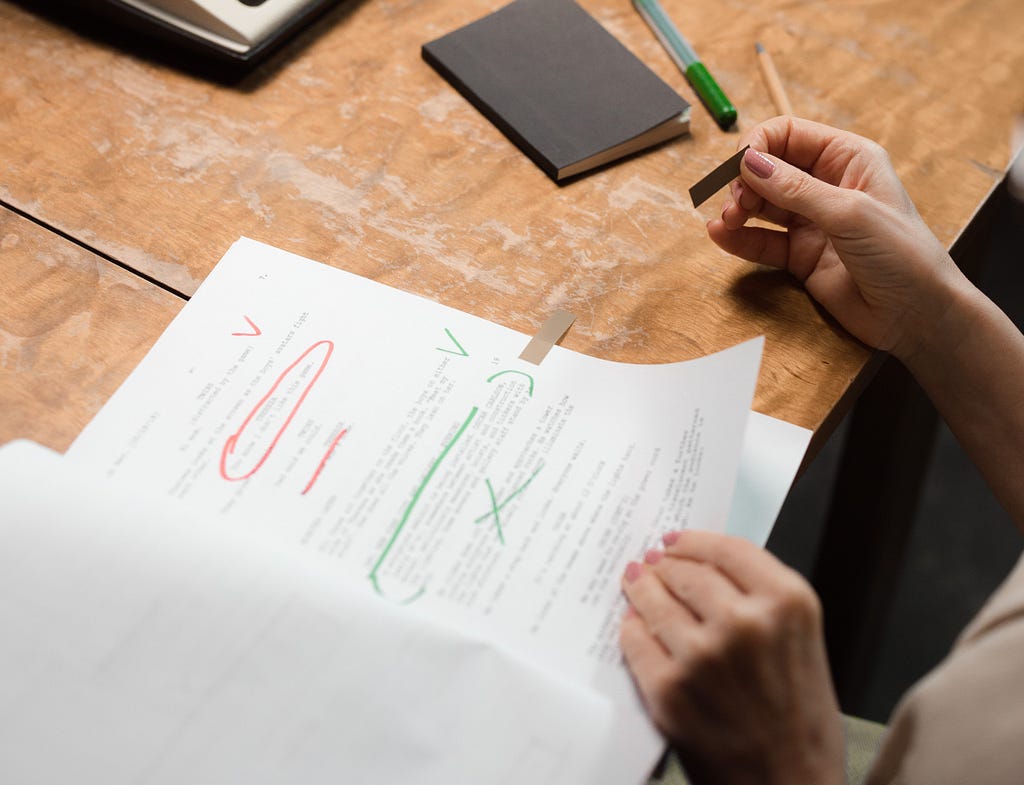 A manuscript on a wooden desk. It has marks in green and red pen on it. A person’s hands are on the manuscript. One hand is about to turn the page and the other is sticking a tab on it. A green pen and small brown notebook are also on the desk.