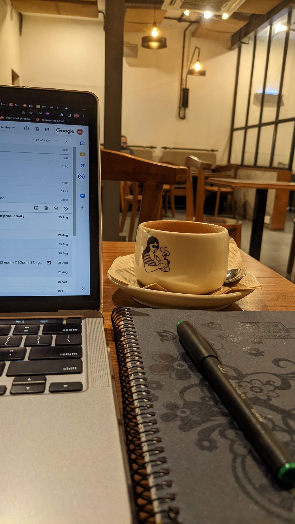 A laptop screen opened at Gmail, flanked by an empty cup of coffee, a spiral bound black notebook, and a capped gel pen, shot in a coffee house