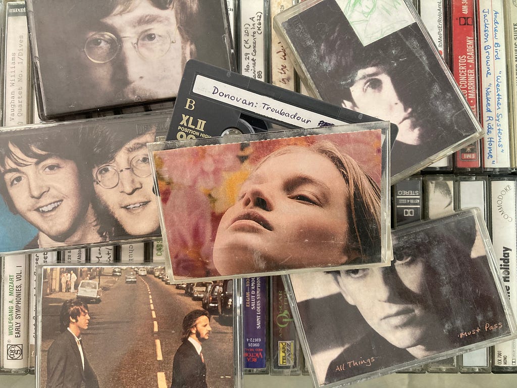 A box of cassette tapes, highlighting Donovan’s “Troubadour” collection (for which I chose an ad of a beautiful woman in front of what appears to be pink and yellow flowers) surrounded by tapes by a few of his good friends, the Beatles. (Photo by author.)