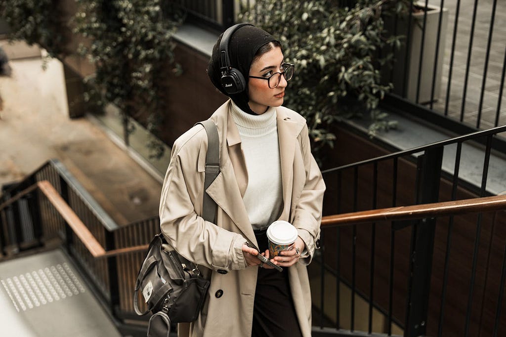 Young Woman in a Beige Coat and Headphones Climbing the Stairs with a Cup of Coffee and a Smartphone in Her Hands | Photo from pexels.com