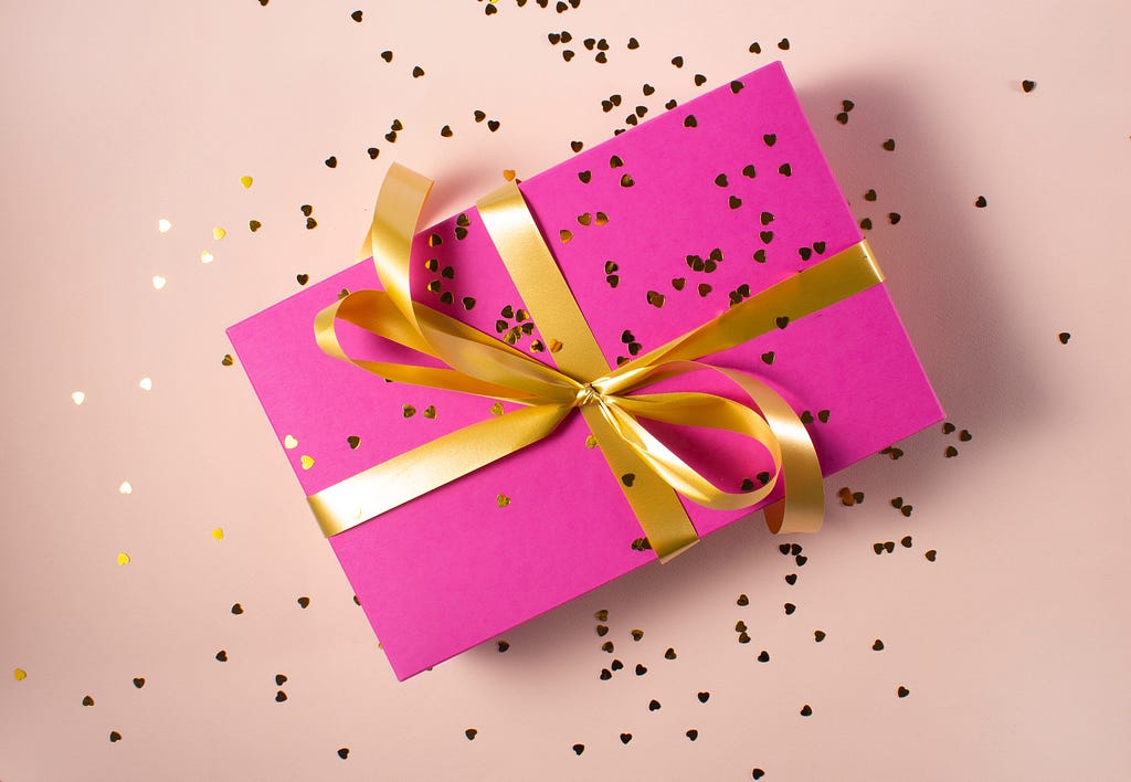 A neon pink box with a gold ribbon and bow on a light pink background with scattered gold confetti.