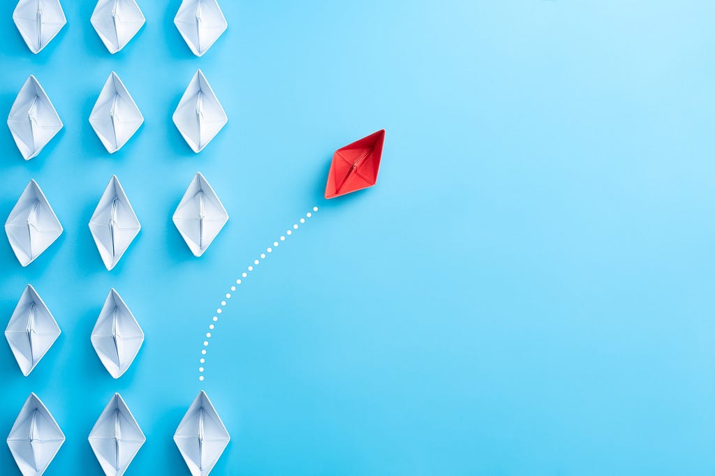 One red paper boat deviating course from a number of white paper boats