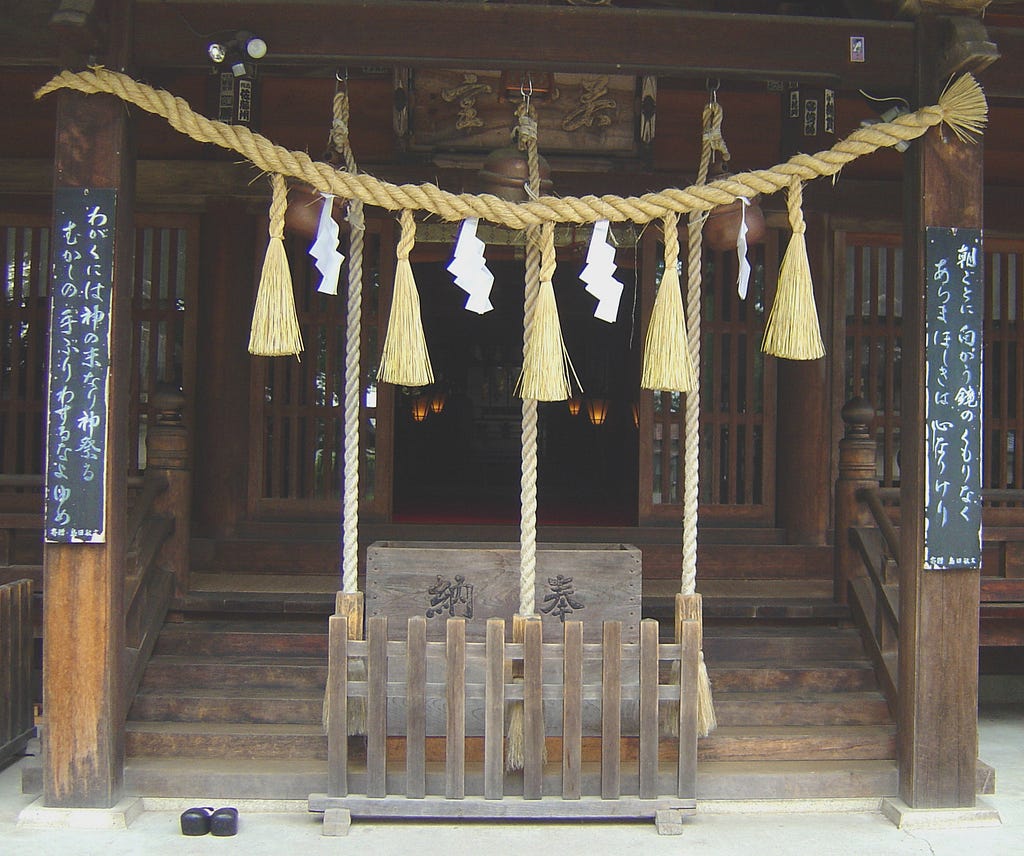 A large wooden box, the size of an old-fashioned travelling trunk, sits below a rope hung with tassels and folded paper gohei at the bottom of a short flight of stairs leading into a dark hall.
