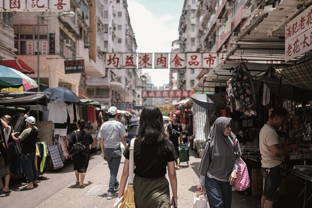 A woman from behind walking through a street with market stalls on the side and Asian characters.