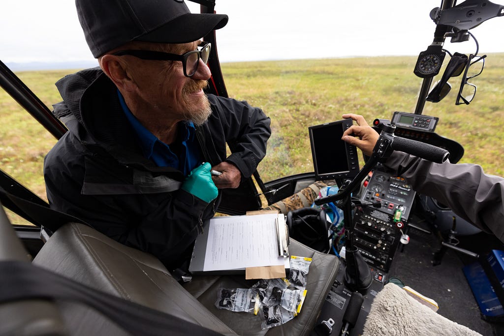A man with a clipboard looks inside a helicopter on a flat grassy plain.