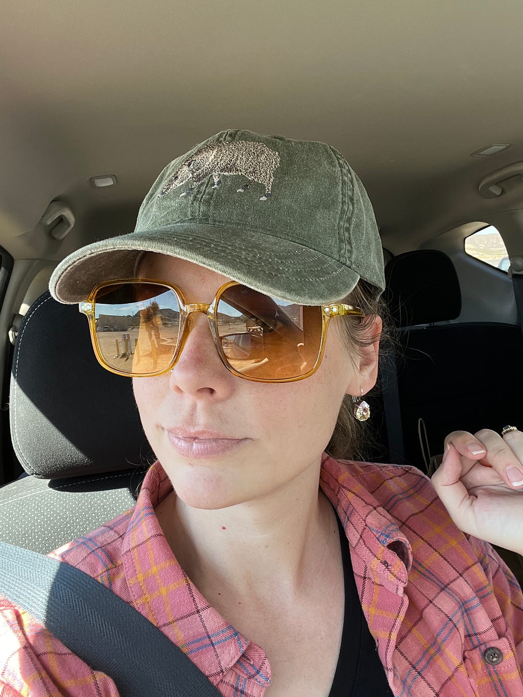 A woman with yellow sunglasses, a pink plaid shirt, and a green hat with a javelina embroidered on it. She has her left hand raised to her shoulder. She is sitting in a car.