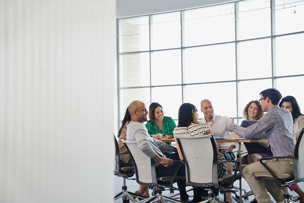 Looking into a team meeting from just outside the room. White wall on the left. Meeting table with 8 team members of varying nationalities discussing while smiling.