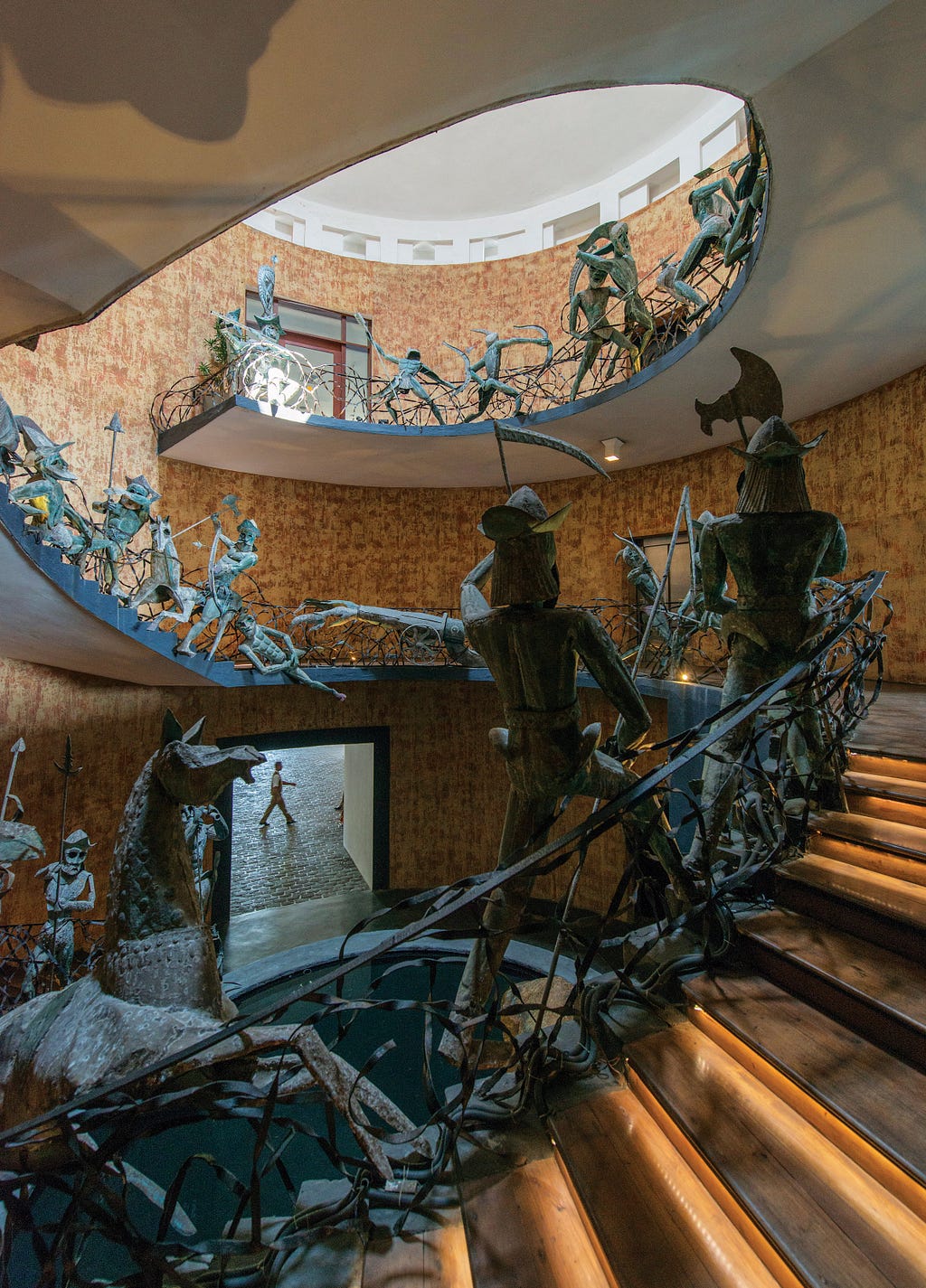 A huge circular staircase made with concrete and the railings are decrorated with metal sculptures of warriors and horses which resembles a battle. The staircase is lit with natural light coming from the sky light at the very top.