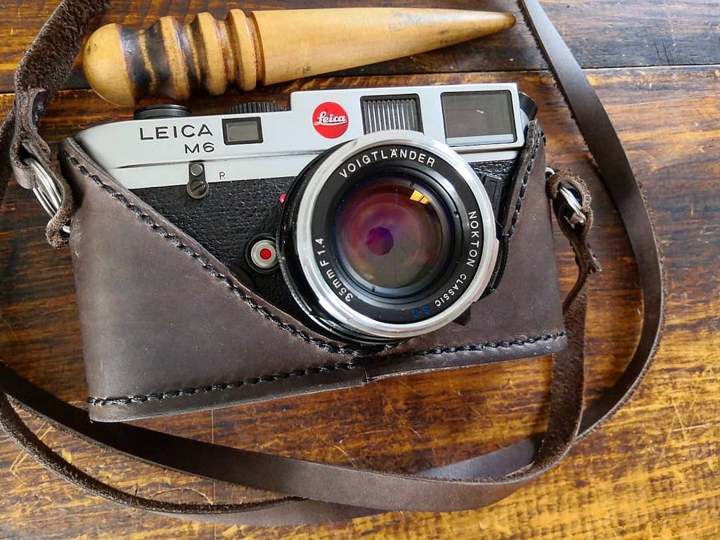 A Leica M6 on a wooden table. The camera has a hand made brown leather half case and matching strap.
