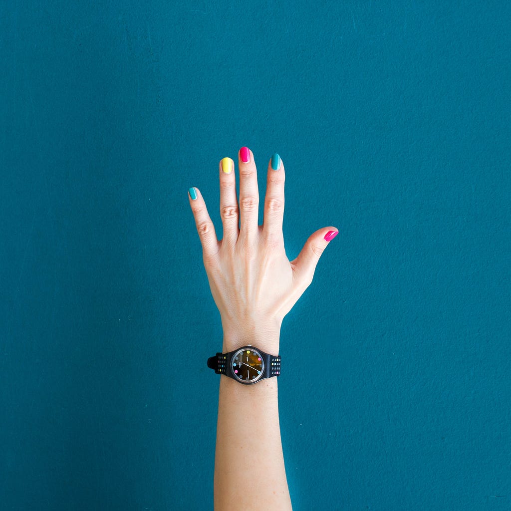 Mão esquerda de uma pessoa, com unhas coloridas e um relógio preto analógico de pulso