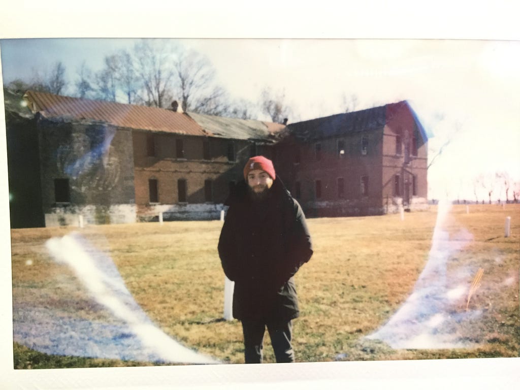 A photograph taken of the author, Michael Agovino, by a wonderful corrections officer. Behind the pillars was the Pheonix Rehab facility, which functioned in the 1970’s. It is crumbling apart and the corrections officer stated that he still finds shoes, clothing, and books in the ruins of the building.