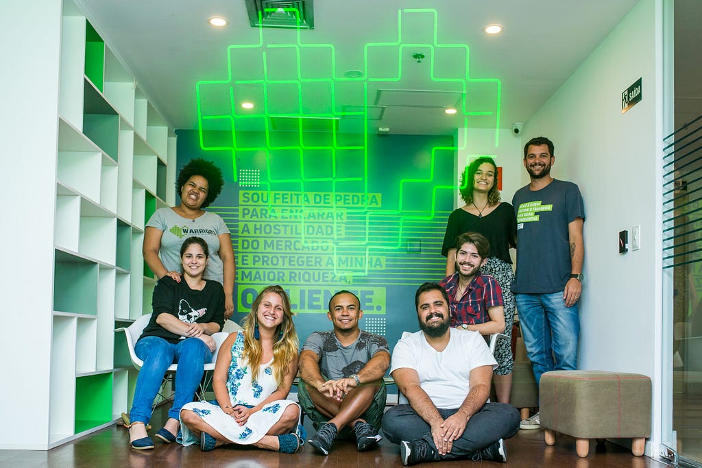 4 pessoas sorrindo para a foto, parte delas sentada e parta delas de pé. Ao meio, uma imagem com um coração verde e luminoso, com desenho similar a pixel art.