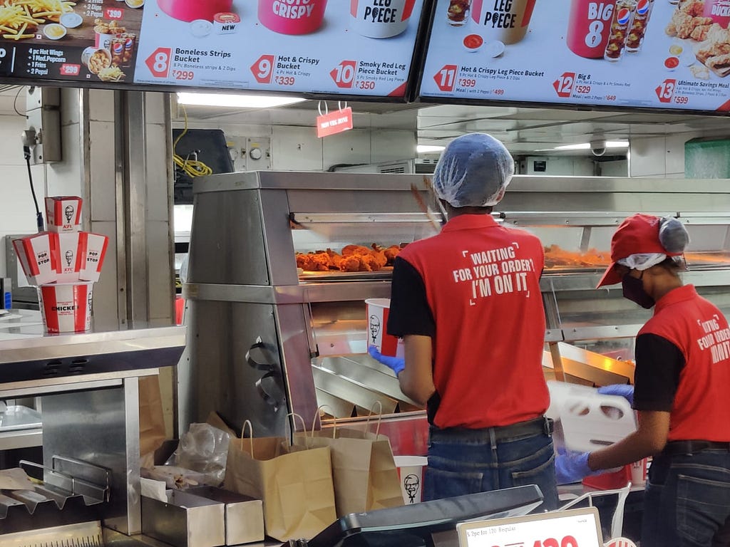 An image where the KFC staff getting the order prepared for the user. On the back of the shirt, it says, waiting for your order? I'm on it.