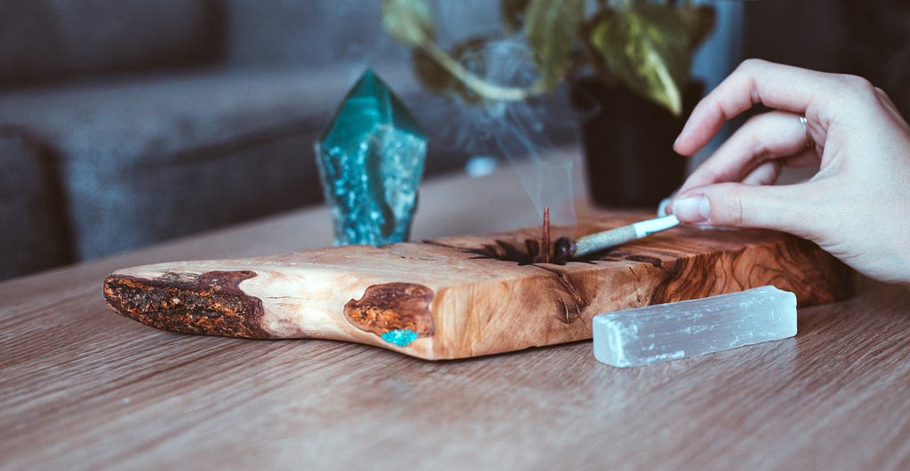 A white woman’s hand ashes a pre-roll hemp joint into a handmade wooden ashtray, decorated with crystals.