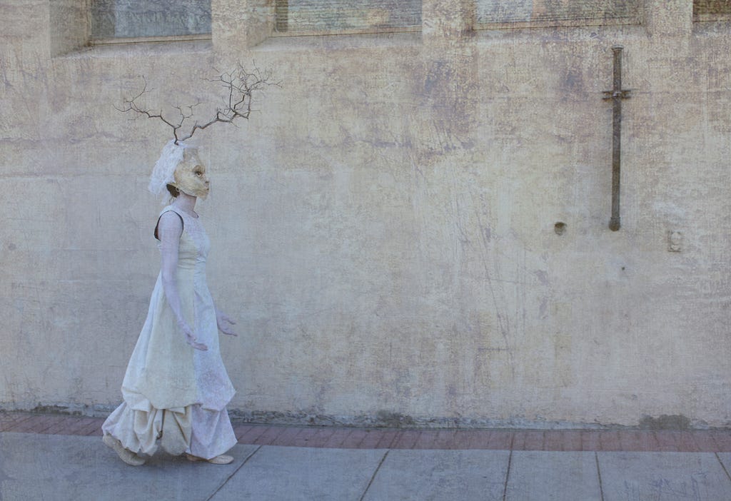 A masked figure in white walk along an old, wall, textured in shades of off-white and beige.