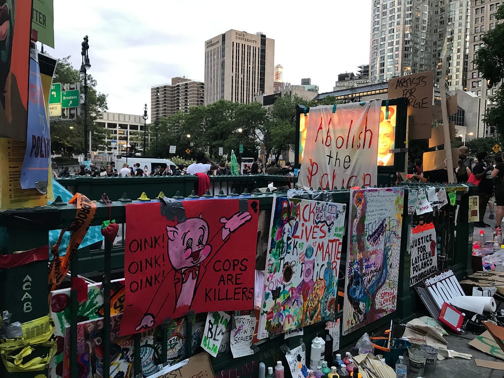 Image is a photograph of a subway station entrance/exit in downtown Manhattan by City Hall covered in hand painted signs about police brutality and abolishing the police. Text on signs includes: “Oink! Oink! Oink! Cops are Killers”, “Abolish the Police”, “Black Lives Matter”, and “Justice for Layleen Polanco”. Further in the distance beyond the subway entrance are a crowd of people gathered. Beyond the people are high rise buildings.