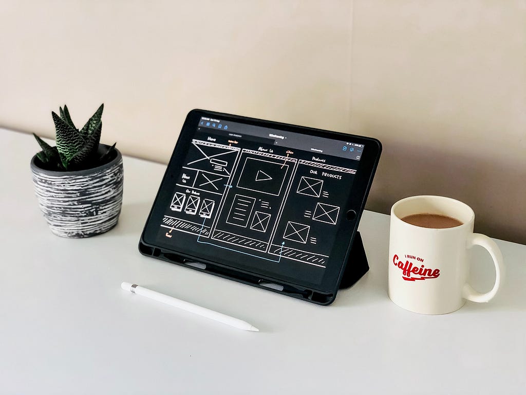 iPad with an example of a minimalist wireframe design with an Apple Pencil in the foreground. An air plant is on the left and a cup of coffee is on the right. The cup says “Caffine”.