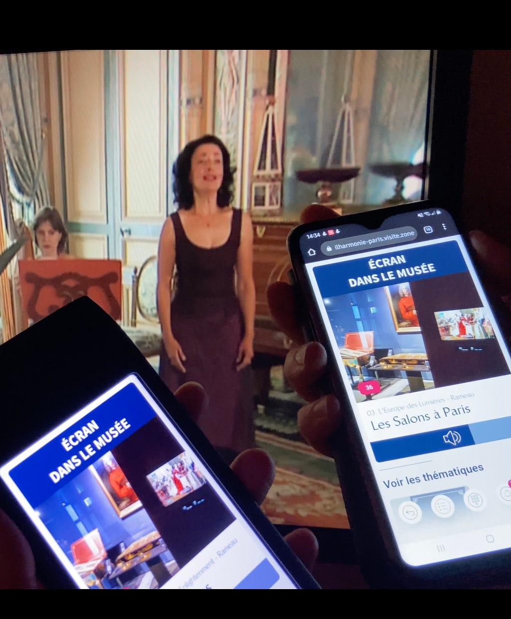 Two smartphones are being held up, displaying content from the museum’s app, and a screen in the back ground shows an opera singer performing. The phones’ screens display the app interface, highlighting ‘Les Salons à Paris’ with options to explore themes.