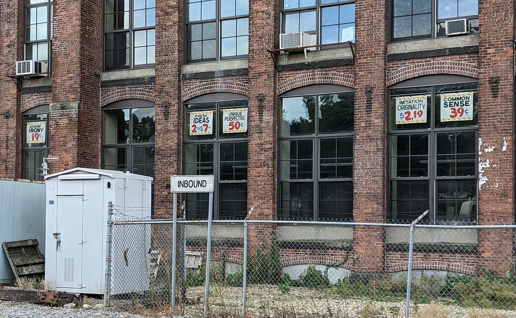 An image of a brick building with large windows. Each window has a sign featuring ironic or unexpected phrases like, “Imitation originality $2.19” or “Unique perspective 50 cents.”