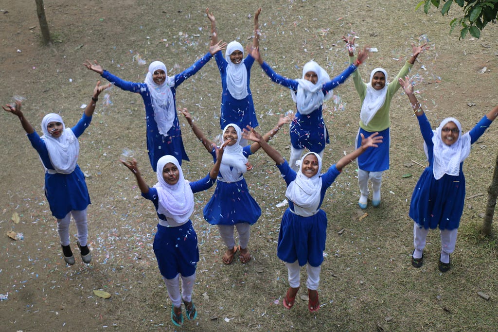 Bangladeshi girls in a blue school uniform and white headscarves throwing up their hands in a celebratory manner with confetti